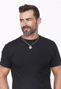 Juoda medvilninė marškinėlių su apvaliu kaklu. Papuošta sidabro spalvos grandinėlės vėriniu su apvaliu pakabuku. Odos atspalvis vidutinis, plaukai tamsiai rudi.