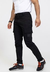 Un pantalon cargo noir avec plusieurs poches, une fermeture à bouton et une coupe fuselée, associé à des baskets noires et orange. Texture de tissu lisse.