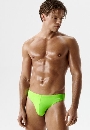 Atletyczny mężczyzna z mokrą skórą, ubrany w jaskrawozielone kąpielówki, stoi na gładkim białym tle, patrząc w dół w lewo.