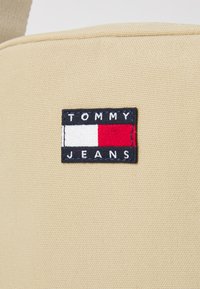 Beige stoffpose med et rektangulært blått felt med "TOMMY JEANS" skrevet i hvit og rød tekst, med et rødt og hvitt rektangulært logo.