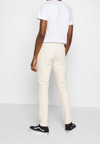 Jasnobeżowe, dopasowane jeansy o gładkiej fakturze z tylnymi kieszeniami, w połączeniu z białym t-shirtem i czarnymi sneakersami, widziane od tyłu.