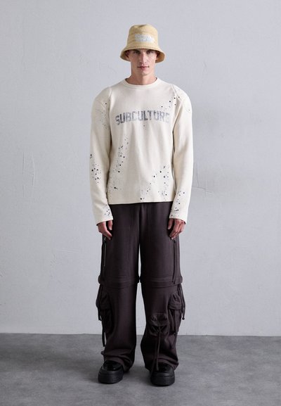 Cream long-sleeve sweater with "SUBCULTURE" text and splatter detail, paired with dark cargo pants featuring zippers and pockets. Black platform shoes.
