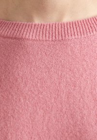 Rožinė megztinė su nertiniu kaklu, pagaminta iš švelnios, lengvos medžiagos. Audinys yra lygus ir vienodas, su subtiliu nertiniu raštu.