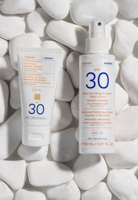 Dwa produkty przeciwsłoneczne na białych kamykach: biały tubka z filtrem przeciwsłonecznym do twarzy w odcieniu oraz biała butelka ze sprayem, oba oznaczone SPF 30.