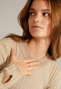Beige ribbed sweater with a wide neckline, featuring a split cuff, worn by a person with light brown hair. A gold ring is worn on their hand.