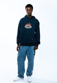 Sudadera azul marino con logo de llamas en rojo y blanco, combinada con pantalones de mezclilla azul claro con parches y un ajuste relajado. Zapatillas blancas.