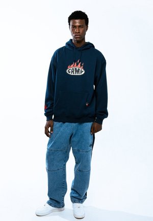 Sudadera azul marino con logo de llamas en rojo y blanco, combinada con pantalones de mezclilla azul claro con parches y un ajuste relajado. Zapatillas blancas.