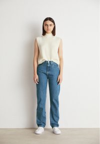 Μπεζ αμάνικο πλεκτό τοπ, με ψηλή λαιμόκοψη; σε συνδυασμό με μπλε ίσια τζιν παντελόνια και λευκά sneakers. Απλός σχεδιασμός, ουδέτερα χρώματα.