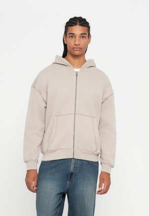 Helle beige Zip-Hoodie mit einer Kordelzugkapuze, großen Vordertaschen, gerippten Bündchen und einem lässigen Schnitt. Trägt man über einem weißen Shirt.