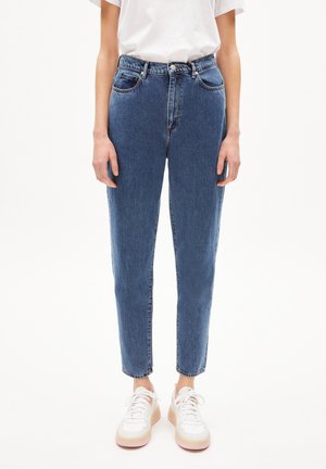 Personne portant un jean bleu taille haute et des baskets blanches, debout devant un fond clair uni.