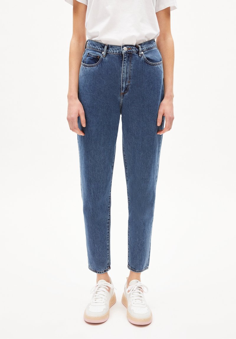 Personne portant un jean bleu taille haute et des baskets blanches, debout devant un fond clair uni.