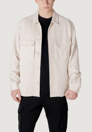 Chaqueta de camisa de manga larga en beige claro, con dos bolsillos en el pecho, botones a presión y un corte relajado, llevada sobre una camiseta negra de cuello redondo.
