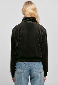 Chaqueta de terciopelo negro con cuello alto y dobladillo acanalado, con mangas largas. Llevada sobre unos jeans de mezclilla azul claros, vista desde la parte posterior.