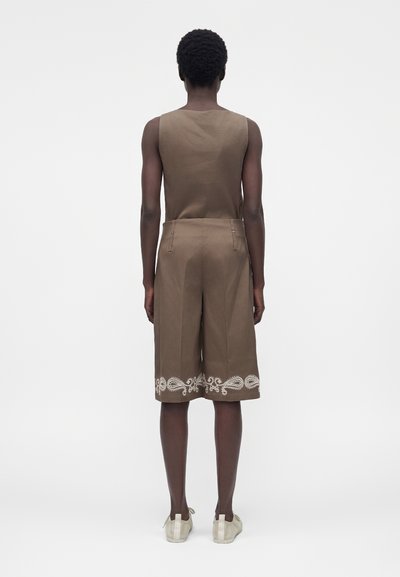 Άτομο στέκεται με την πλάτη προς τα έξω, φοράει αμάνικη καφέ μπλούζα και αντίστοιχα σορτς μέχρι το γόνατο με λευκό διακοσμητικό μοτίβο κοντά στα τελειώματα, λευκά παπούτσια.