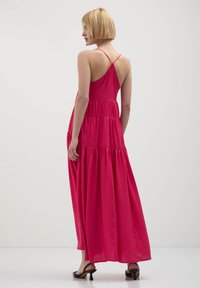 Donna con capelli corti biondi, che indossa un vestito lungo senza maniche, incrociato sul retro, di un vibrante rosa fucsia e sandali neri con tacco, in piedi di profilo.