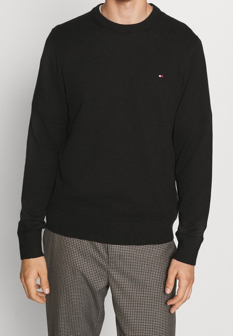 Maglione nero in maglia con scollo rotondo, polsini e orlo a costine. Presenta un piccolo logo rosso e bianco sul petto. Indossato con pantaloni a quadri.