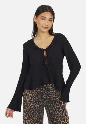 Vrouw die een zwarte top met lange mouwen draagt met strikjes en ruches, gecombineerd met een broek met luipaardprint, staand tegen een effen achtergrond.