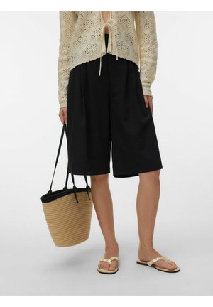 Osoba nosiaca čierne široké šortky po kolená, béžový háčkovaný top, sandále zdobené perlami, držiaca svetlohnedú pletenú kabelku s čiernymi rukoväťami.