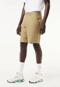 Pantaloni shorts beige con un design sartorial, dotati di chiusura a bottone e tasche frontali, abbinati a scarpe da ginnastica bianche e calze.