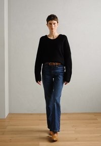 Black, long-sleeve sweater; blue flared jeans; brown belt; tan heeled boots; standing on wooden floor against a light gray wall.