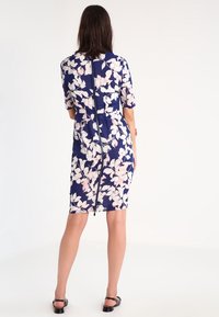 Blommig knälång klänning i marinblått med vita och rosa blommor. Har korta ärmar och en central bakdragkedja. Slät tygtextur.