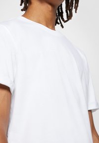 Biała bawełniana koszulka z okrągłym dekoltem i krótkimi podwiniętymi rękawami. Miękka tekstura i luźny krój, z widocznymi detalami szycia na kołnierzu.