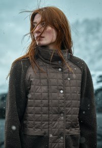 Bruine gewatteerde jas met een hoge kraag, voorzien van een textuur fleece lichaam en drukknoop details aan de voorkant. Sneeuwvlokken zichtbaar.