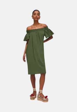 Donna che indossa un vestito verde oliva con spalle scoperte fino al ginocchio e sandali platform beige con accenti di fiori rossi, in piedi su sfondo bianco.
