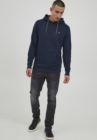 Marineblauer Hoodie mit einer Fronttasche, verstellbaren Kordeln und einem kleinen Logodetail. Kombiniert mit dunklen Jeans und schwarzen Sneakers.