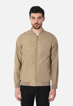 Uomo che indossa una giacca bomber beige con cerniera, polsini e collo a costine, in piedi davanti a uno sfondo chiaro e uniforme.