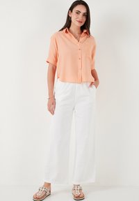 Short-sleeve, button-up shirt in light orange fabric, paired with wide-leg white pants and sandals with embellishments.