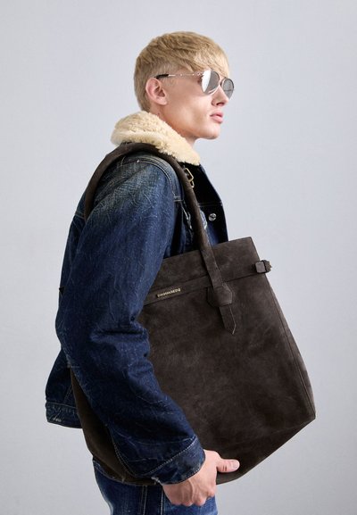 Blondais vīrietis ar aviatora brillēm un džinsa jaku ar vilnas apkakli, nes liela brūna zamša Dsquared2 rokassomu.