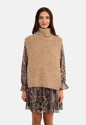 Donna che indossa un maglione beige lavorato a maglia senza maniche con collo alto sopra un vestito a maniche lunghe con stampa animalier, in piedi davanti a uno sfondo bianco semplice.