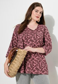 Blusa rosa con un motivo floreale e scollo a V, abbinata a una borsa di paglia intrecciata con manici in pelle. I jeans grigi completano l'outfit.