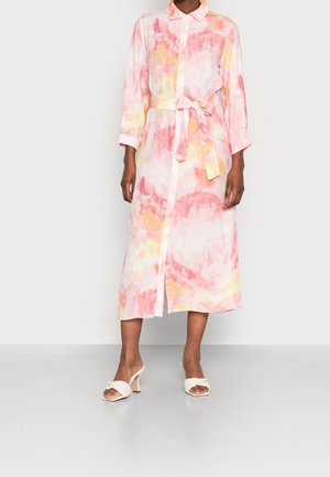 Mujer que lleva un vestido de manga larga, abotonado, con estampado acuarela en rosa y amarillo, con cinturón anudado en la cintura y sandalias blancas de tacón.