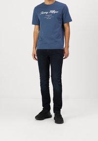 Blå crew-neck t-shirt med vitt logotryck, kombinerad med mörka denimjeans med slitna detaljer och svarta träningsskor.