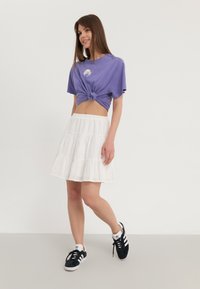 Lila, geknotetes Baumwoll-T-Shirt mit Grafik, kombiniert mit einem weißen, gestuften, strukturierten Rock. Schwarze und weiße Sportschuhe runden das Outfit ab.