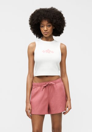 Fiatal nő göndör hajjal, fehér ujjatlan crop topot visel, amelyen rózsaszín virágmintás, valamint rózsaszín húzózsinóros rövidnadrágot, egy egyszerű háttér előtt áll.