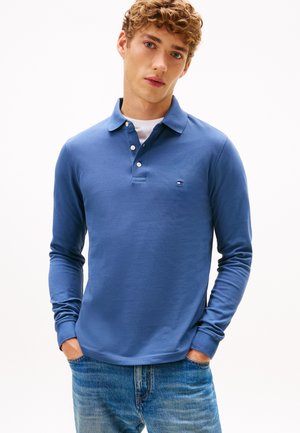 Νέος άνδρας με σγουρά μαλλιά που φοράει μπλε πόλο με μακριά μανίκια πάνω από λευκή φανέλα, ντυμένος με μπλε τζιν, χέρια στις τσέπες.