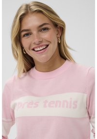 Mujer sonriente con cabello rubio que lleva pendientes de aro dorados y un suéter rosa con una franja blanca y el texto "après tennis".