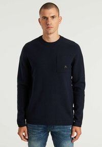 Maglietta a maniche lunghe blu navy con colletto a girocollo, dotata di tasca frontale e dettagli di cucitura sottili, abbinata a jeans azzurri sbiaditi.