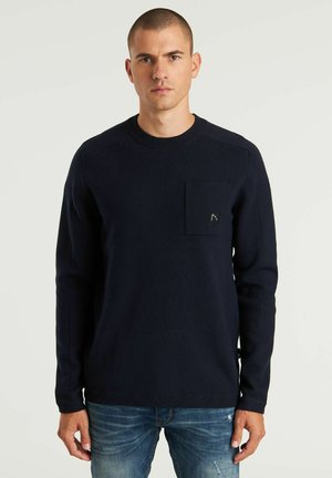 Marineblaues Langarmshirt mit Rundhalsausschnitt, ausgestattet mit einer Brusttasche und dezenten Steppdetails, kombiniert mit hellblauen, schlichten Jeans.