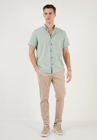 Cămașă cu mânecă scurtă, dintr-un material verde deschis, cu guler și nasturi negri. Asortată cu pantaloni slim-fit bej deschis și adidași bej.