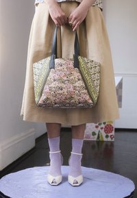 Osoba ubrana w beżową spódnicę i liliowe skarpety, nosząca białe sandały na obcasie, trzyma torbę na ramię w kwiatowy wzór z czarnymi uchwytami.