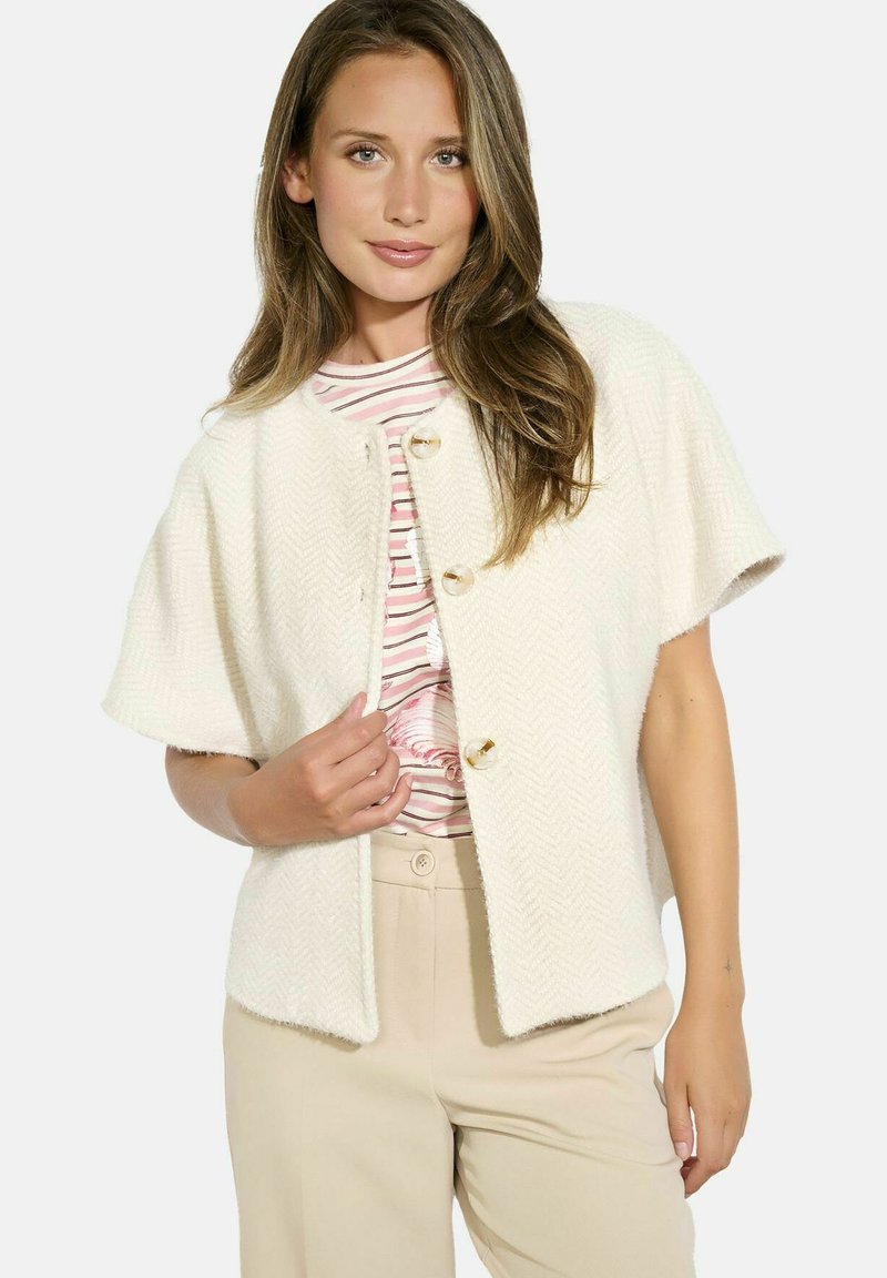 Cream herringbone-patterned cardigan with short sleeves, featuring gold-button fastenings. Worn over a striped shirt and beige trousers.