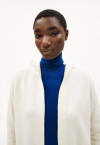 Weißer Kapuzenkarten aus strukturiertem Stoff, getragen über einem einfarbigen blauen Rollkragenpullover. Fokus auf die klaren Linien und das sanfte Erscheinungsbild des Kleidungsstücks.