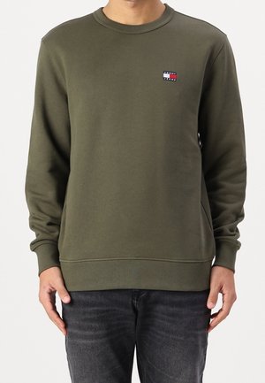 Mężczyzna ubrany w oliwkowo-zieloną bluzę z długim rękawem i okrągłym dekoltem z małym logo Tommy Jeans na piersi, w połączeniu z ciemnoszarymi dżinsami.