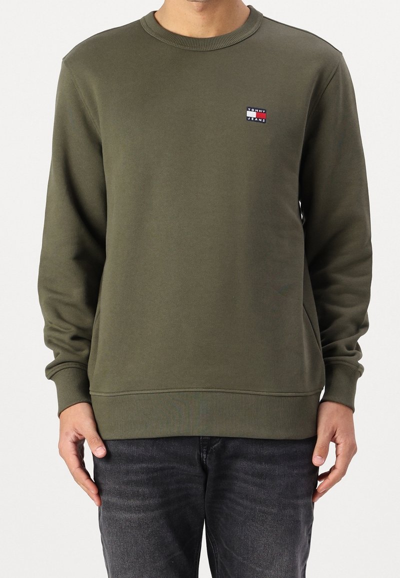 Homme portant un sweat-shirt à col rond olive à manches longues avec un petit logo Tommy Jeans sur la poitrine, associé à un jean gris foncé.