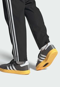 Baskets grises avec des rayures blanches, semelle jaune et tige en tissu gris. Associées à un pantalon noir avec des rayures blanches sur les côtés.