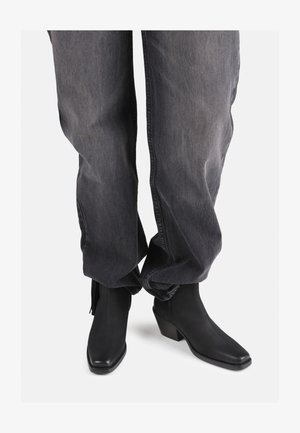 Czarne skórzane botki za kostkę z wydłużonym noskiem i niskim, kwadratowym obcasem, zestawione z luźnymi, wyblakłymi, szarymi dżinsami.